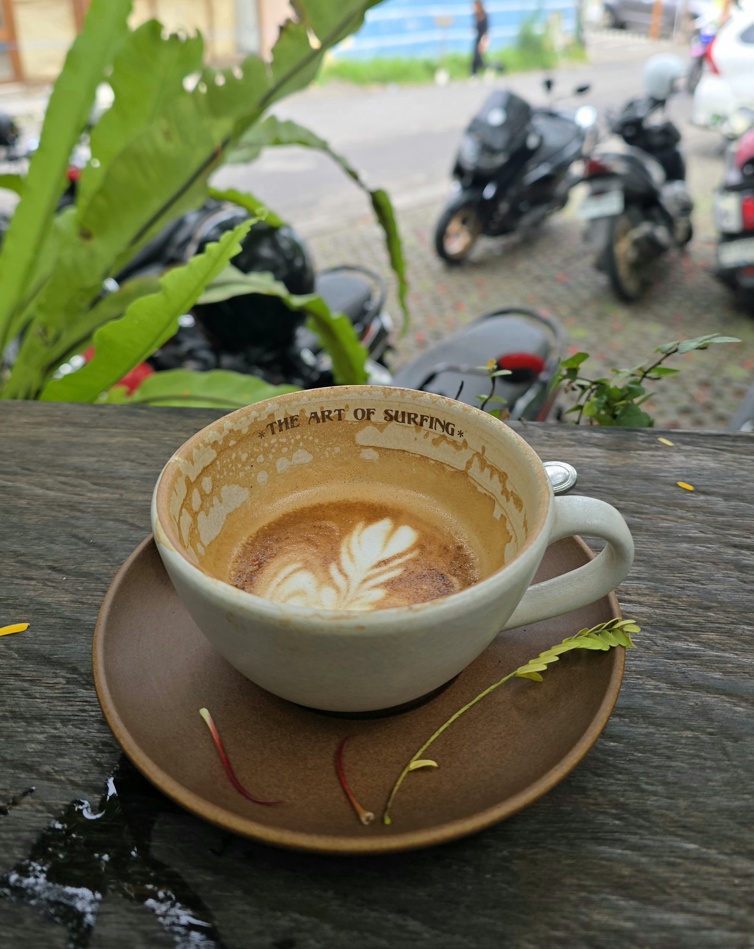A latte in a ceramic cup reading The Art of Surfing, on a weathered wooden table with motorbikes parked behind, Bali